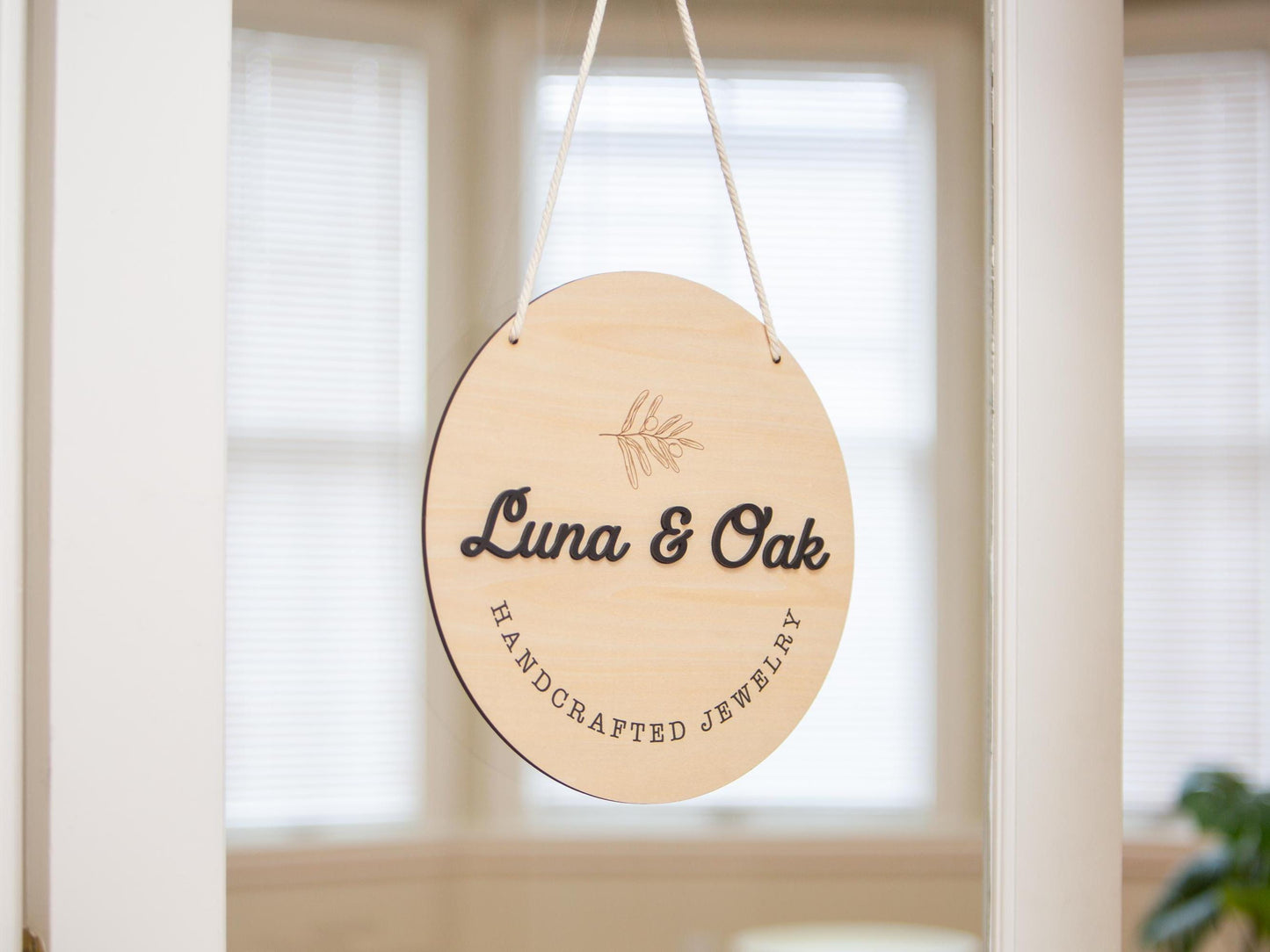 May include: A round, wooden sign with the text &#39;Luna & Oak Handcrafted Jewelry&#39; in black lettering. The sign is hanging from a white string and has a small, engraved leaf design in the center.