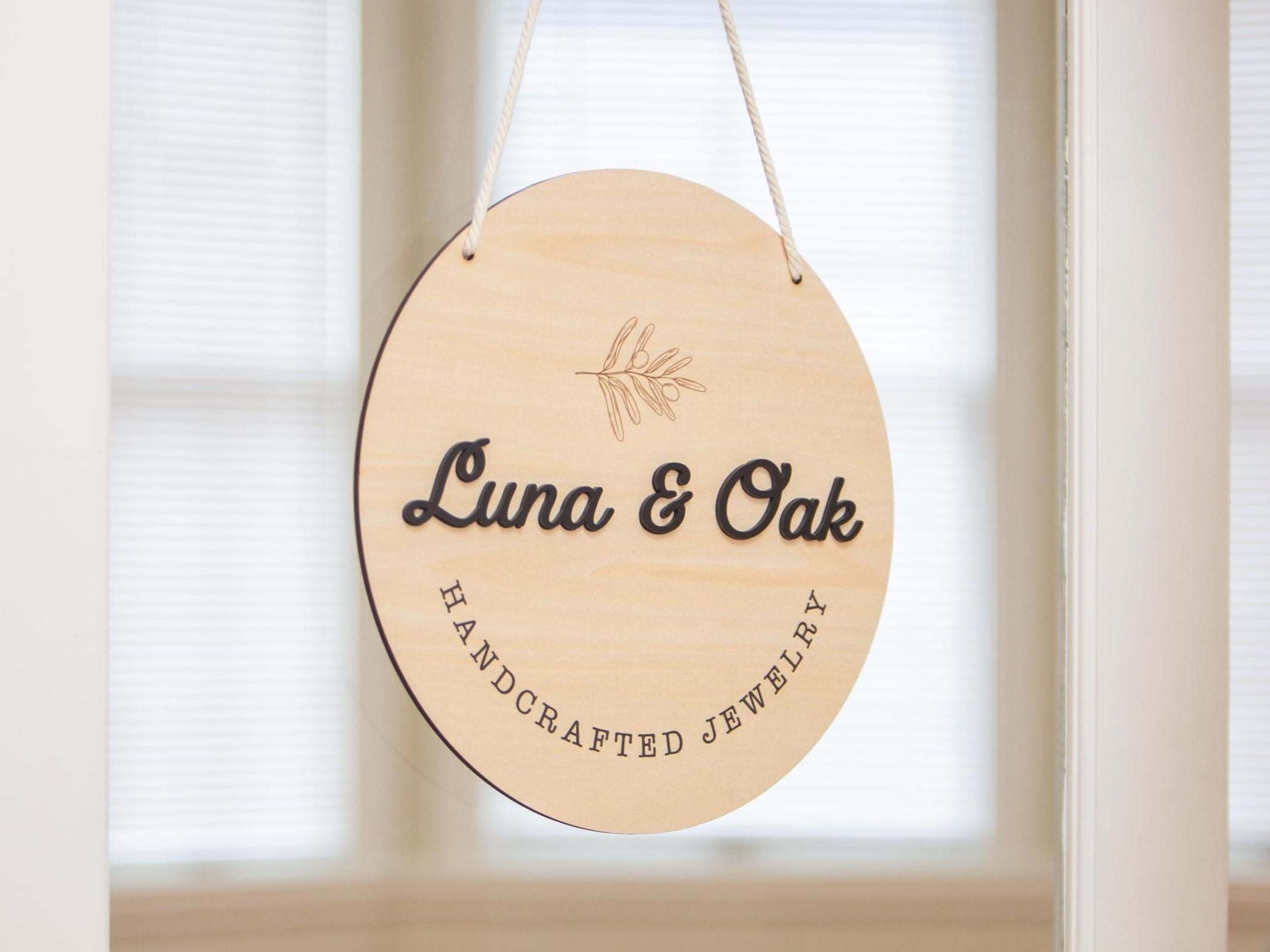 May include: A round, wooden sign with the text 'Luna & Oak Handcrafted Jewelry' in black lettering. The sign is hanging from a white string and has a small, engraved leaf design in the center.