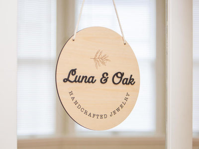 May include: A round, wooden sign with the text &#39;Luna & Oak Handcrafted Jewelry&#39; in black lettering. The sign is hanging from a white string and has a small, engraved leaf design in the center.