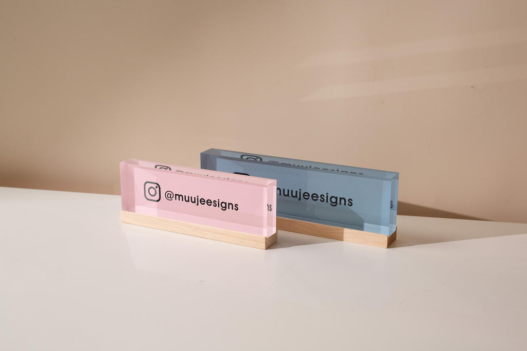 May include: Two rectangular acrylic business card holders. One is pink, the other blue. Each has a wooden base and displays the Instagram handle '@muujeedesigns' along with the Instagram logo. The pink holder is on the left, the blue on the right, both against a neutral background.