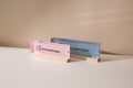 May include: Two rectangular acrylic business card holders. One is pink, the other blue. Each has a wooden base and displays the Instagram handle '@muujeedesigns' along with the Instagram logo. The pink holder is on the left, the blue on the right, both against a neutral background.