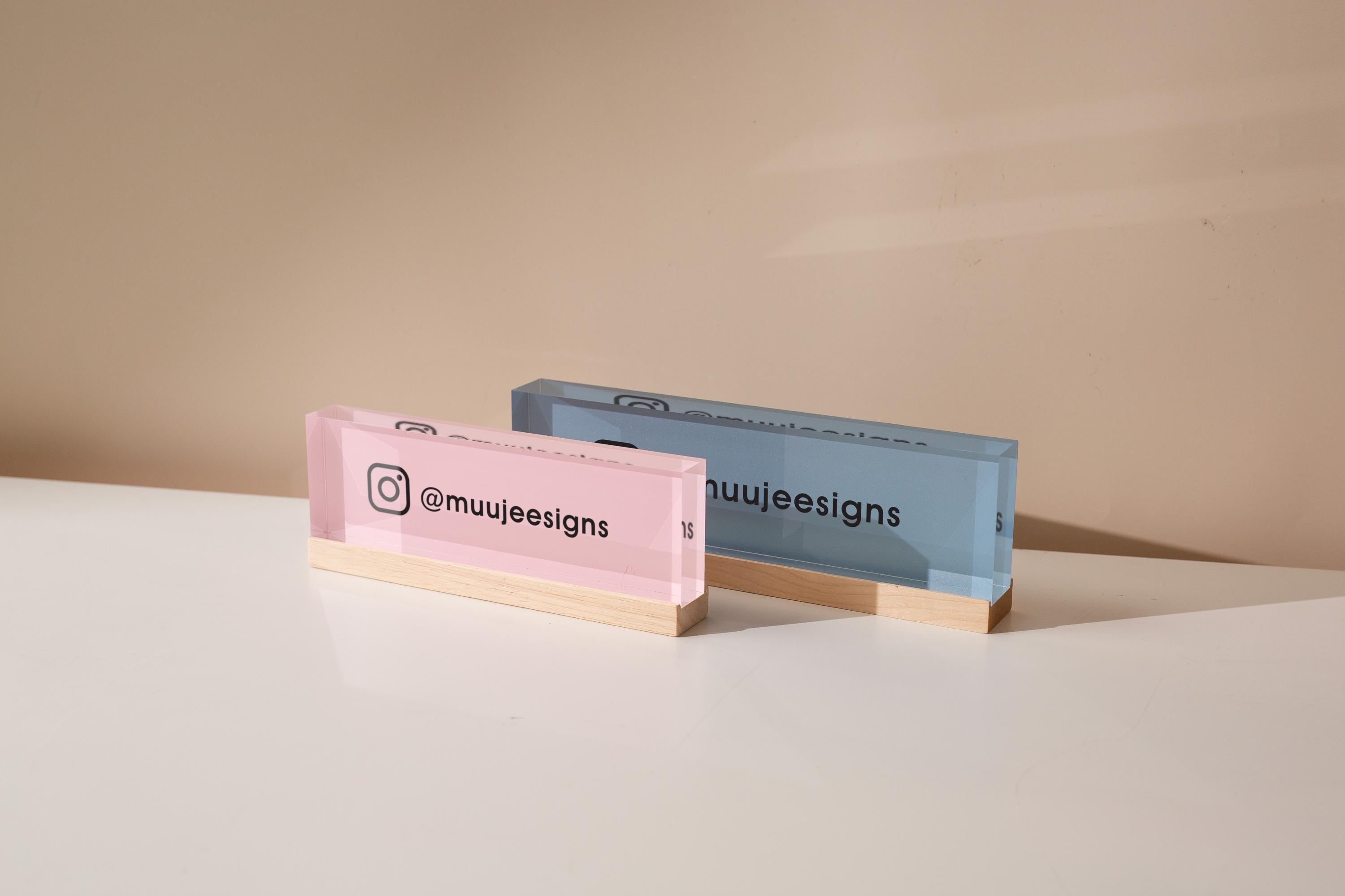May include: Two rectangular acrylic business card holders. One is pink, the other blue. Each has a wooden base and displays the Instagram handle '@muujeedesigns' along with the Instagram logo. The pink holder is on the left, the blue on the right, both against a neutral background.