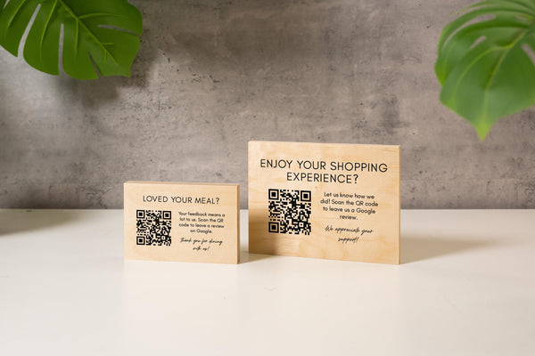 May include: Two wooden signs with printed text and QR codes. One sign reads "LOVED YOUR MEAL?" and the other says "ENJOY YOUR SHOPPING EXPERIENCE?". Both signs have a light wood grain finish and are on a white surface.